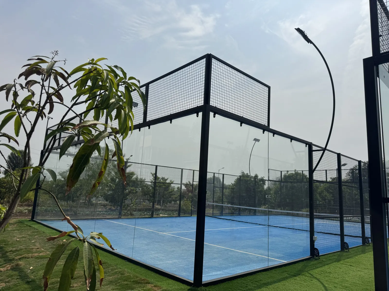 Outdoor padel court with a blue playing surface, glass walls and a black steel frame, set on artificial turf